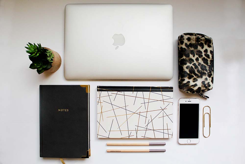 a spread of beautiful objects carefully placed: a mac laptop, iphone, a plant, two notebooks, and two pens