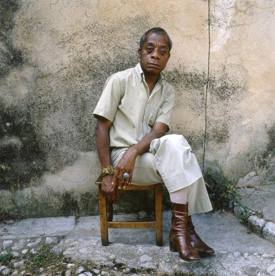 James Baldwin, sitting sideways in a chair, looking soulful.